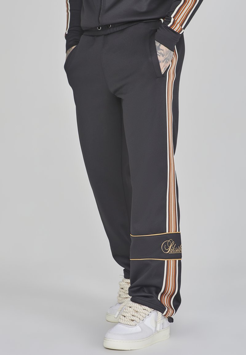 Zwarte sportbroeken met verticale strepen in oranje, wit en bruin, met zijzakken en een gouden geborduurd logo bij de zoom.