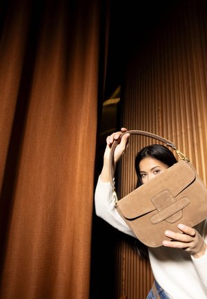 Brown suede handbag with a flat, structured design and a stitched flap. Features a top handle and a gold hardware accent.