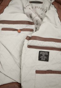 Braunleder-aczentierte Innenseite einer Jacke, die mit einem leichten, gesteppten Futter, mehreren Taschen und einem Etikett, das die verwendeten Materialien beschreibt, ausgestattet ist.