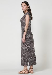 Braun gemustertes Maxikleid mit kurzen Ärmeln, das eine Mischung aus Kreisen und Linien in helleren Farbtönen zeigt, mit Beinschlitzen und kombiniert mit braunen Sandalen.