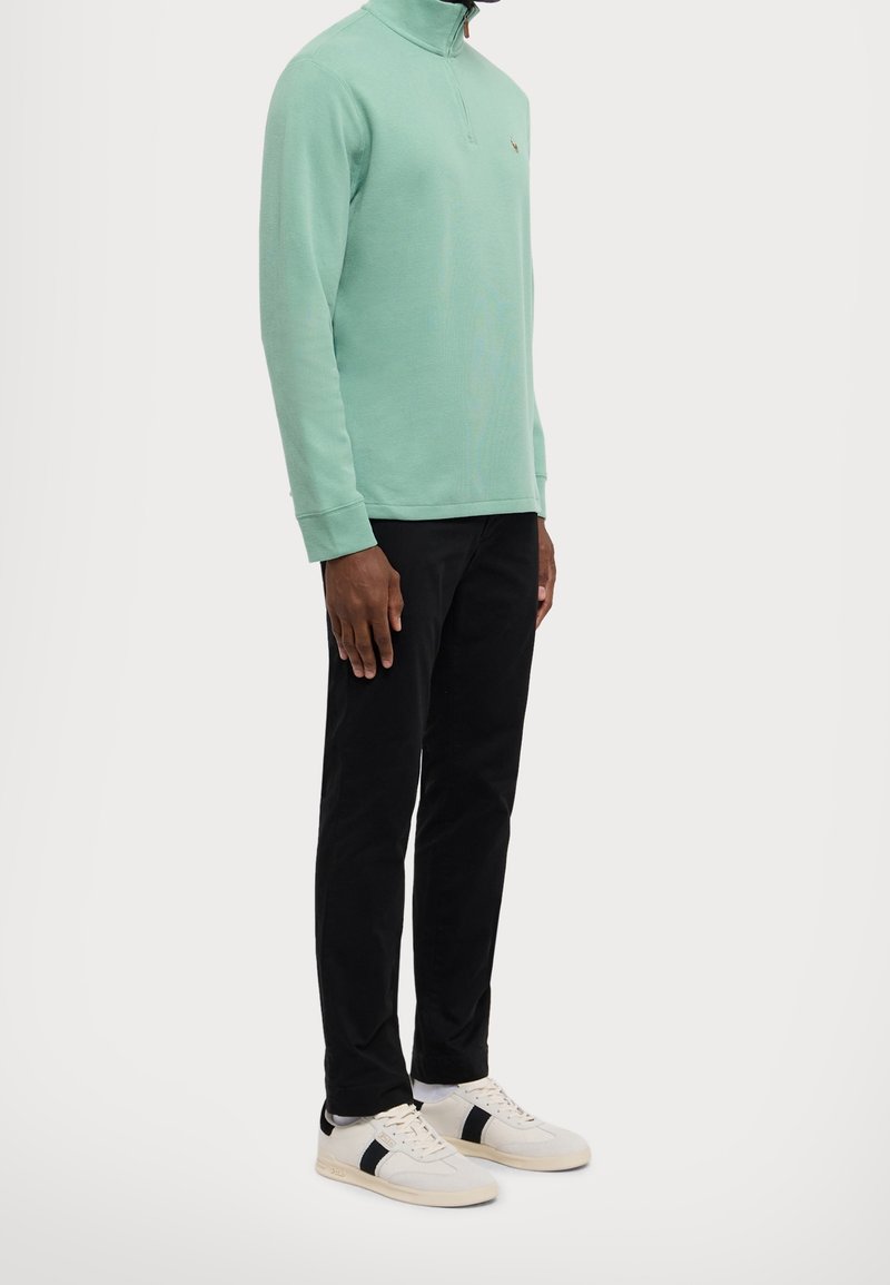 Personne portant un pull vert menthe à fermeture éclair quart de zip, pantalon noir et baskets blanches avec des rayures noires, debout devant un fond uni.