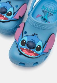 Blå träskor gjorda av gummi, med en upphöjd design av ett tecknat karaktärs ansikte med stora öron, leende uttryck och livfulla färger.