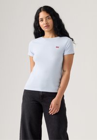 Světle modré tričko s krátkým rukávem vyrobené z měkké látky, které má malé červené logo Levi's na levé hrudi a přiléhající střih. Černé kalhoty.