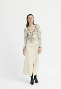 Langärmliges Strickoberteil mit tiefem V-Ausschnitt und schwarzem Saum, kombiniert mit einem beigefarbenen Maxirock mit seitlichem Schlitz und Schnürdetails. Schwarze Schuhe.