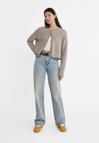 Beige knit cardigan with round neckline and button front, worn over a white crop top. Pair of light blue wide-leg jeans and yellow sneakers.