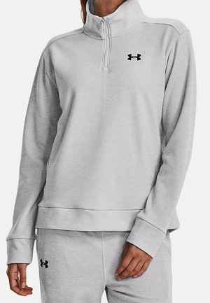 Osoba ubrana w jasnoszary sweatshirt z zamkiem do połowy i dopasowane spodnie z czarnymi logo Under Armour na piersi i udzie.