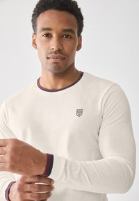 Camisa blanca de manga larga con cuello redondo, que presenta un ribete a rayas en color burdeos y azul marino y un pequeño logo bordado en el pecho.