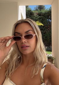 Hosszú szőke hajú nő apró rózsaszín napszemüveget igazít, bézs fal mellett áll, háttérben zöld növényzet és kék ég látható.