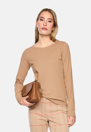 Woman wearing beige long-sleeve shirt and plaid pants holding a brown leather handbag with gold chain detail against a plain background.