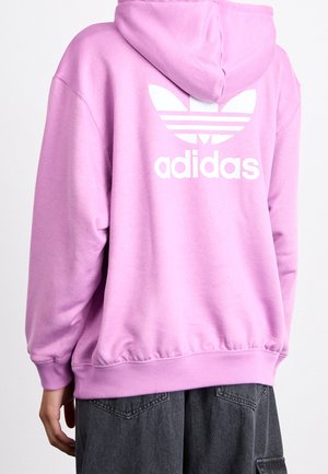 Persoon draagt een lichtpaarse Adidas-hoodie met een wit trefoil-logo en tekst op de achterkant, gecombineerd met donkergrijze broek.