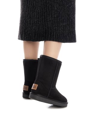 Botas de tobillo de ante negro con punta redonda y suela de goma. Presentan detalles de costura y un parche de logo marrón en la parte trasera.
