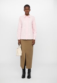 Camicia button-up rosa chiaro con ricami floreali, abbinata a una lunga gonna marrone con uno spacco frontale. Stivali neri al ginocchio e una borsa bianca.