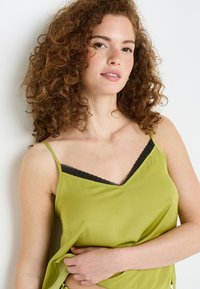 Top canotta verde realizzato in tessuto liscio, con spalline sottili e una finitura in pizzo nero lungo il scollo. Design semplice e vestibilità comoda.
