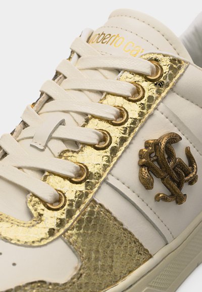 Baskets blanches avec accents texturés dorés, lacets beiges, œillets métalliques et logo décoratif complexe en bronze sur le panneau latéral.