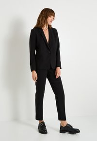 Traje negro a medida que presenta un blazer de escote en V profundo con un solo botón, pantalones ajustados y zapatos negros con suelas gruesas.