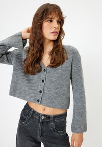Grijze cropped cardigan, geribbelde textuur, V-hals, zwarte knopen, brede mouwen. Gecombineerd met hoge zwarte jeans. Minimalistisch ontwerp.