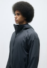 Veste coupe-vent noire avec un col montant, fermeture éclair et poches latérales. Légère, texture lisse et coupe décontractée.