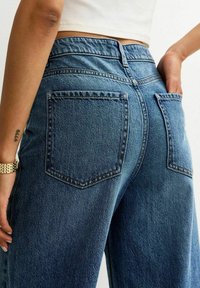 Persona que lleva puestos unos jeans de mezclilla azul de tiro alto con bolsillos traseros, una blusa corta blanca, un reloj de oro en la muñeca y un tatuaje en el antebrazo izquierdo, con la mano en el bolsillo.