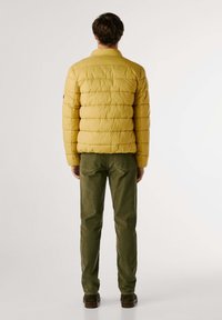 Chaqueta de plumas amarilla con costuras horizontales, mangas largas y cuello alto, combinada con pantalones verde oliva y zapatos marrones.