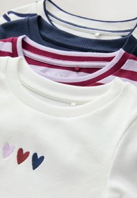 Tri t-shirtova na hrpi: bijela rebrasta majica s izvezanim srcima, mornarskoplava prugasta majica i bordo prugasta majica s svjetlijim prugama.