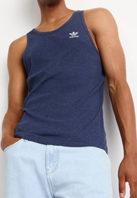 Top de tirantes azul con acanalado y escote redondo, que presenta un pequeño logo blanco de Adidas en el lado izquierdo. Combinado con pantalones de mezclilla claros.