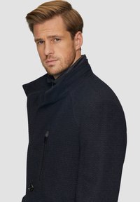 Marineblauwe wollen blend jas met een gestructureerd oppervlak, hoge kraag en een ritssluiting op de borstzak. Voorzien van grote knopen en gestructureerde schouders.