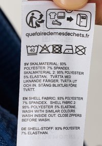 Etykieta pielęgnacyjna odzieży z zawartością materiału: 93% poliester, 7% spandex. Wiele języków, instrukcje prania i symbole pielęgnacyjne nadrukowane na białym materiale.