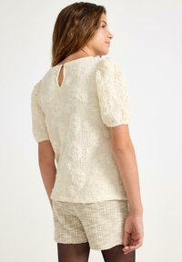 Kerman tekstuuri blouse, jossa on puffi hihat, kukkakuosi ja takana avainaukko, yhdistettynä kevyisiin vaaleisiin shortseihin.