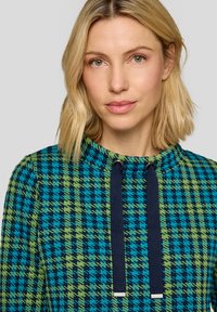 Kapuzenpullover in Blau-, Grün- und Marine-Hahnentrittmuster mit strukturierter Oberfläche. Verfügt über eine Kordel am Kragen mit metallischen Details.