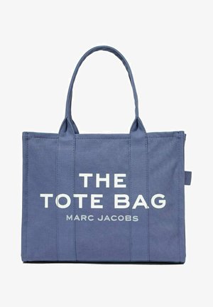 Blå lerret tote bag med hvit tekst som sier "THE TOTE BAG MARC JACOBS," med to robuste håndtak og rektangulær form.