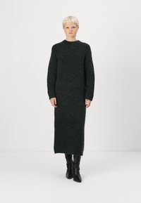 Πλεκτό φόρεμα σε σκούρο πράσινο, με μακριά μανίκια και ψηλό λαιμό, υφή υφάσματος, ίσια σιλουέτα, φτάνει μέχρι το μέσο της γάμπας, συνδυασμένο με μαύρες μπότες μέχρι το γόνατο.