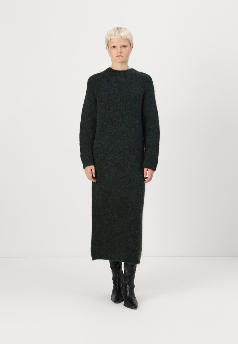 Πλεκτό φόρεμα σε σκούρο πράσινο, με μακριά μανίκια και ψηλό λαιμό, υφή υφάσματος, ίσια σιλουέτα, φτάνει μέχρι το μέσο της γάμπας, συνδυασμένο με μαύρες μπότες μέχρι το γόνατο.