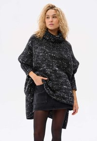 Czarny i biały nakrapiany wełniany ponczo z wysokim dekoltem, w oversize'owym kroju, z bocznymi kieszeniami, nałożone na czarną spódnicę i rajstopy.