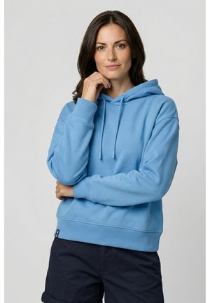 Frau mit halblangem dunklem Haar, die einen hellblauen Hoodie und dunkle Hosen trägt, posiert mit einer Hand nahe ihrem Kinn vor einem einfarbigen Hintergrund.