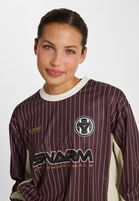 Kvinde med brunt hår og grønne øjne iført en vinrød sportsdragt med tynde striber med "SWARM" og cirkulært logo på brystet, smilende blidt.