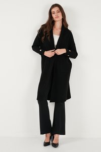 Svart ribbad cardigan med öppen front, långa ärmar, fickor och detaljer i guldtonad knappar, kombinerad med svarta byxor och klackar.