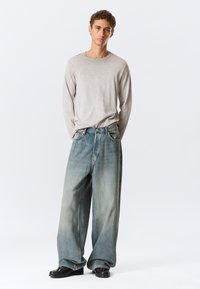 Ανοιχτό γκρι πουκάμισο με μακριά μανίκια συνδυασμένο με φαρδιά, ξεθωριασμένα μπλε τζιν με βαθμιαίο εφέ ομπρέ και σκούρα παπούτσια.