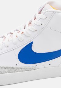 Bílé kožené tenisky s modrým logem Swoosh, texturovanou šedou špičkou, polstrovaným jazykem a bílými tkaničkami na jednobarevném pozadí.