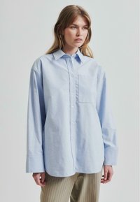 Cămașă albastru deschis, cu mâneci lungi și nasturi, fabricată din bumbac. Are un buzunar pe piept, hem rotunjit și o croială relaxată. Este combinată cu pantaloni bej cu dungi.
