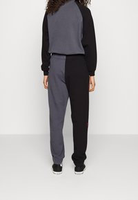 Persona con pantalones de chándal gris y negro a dos tonos y sudadera a juego, de pie con la espalda hacia la cámara contra un fondo plano.