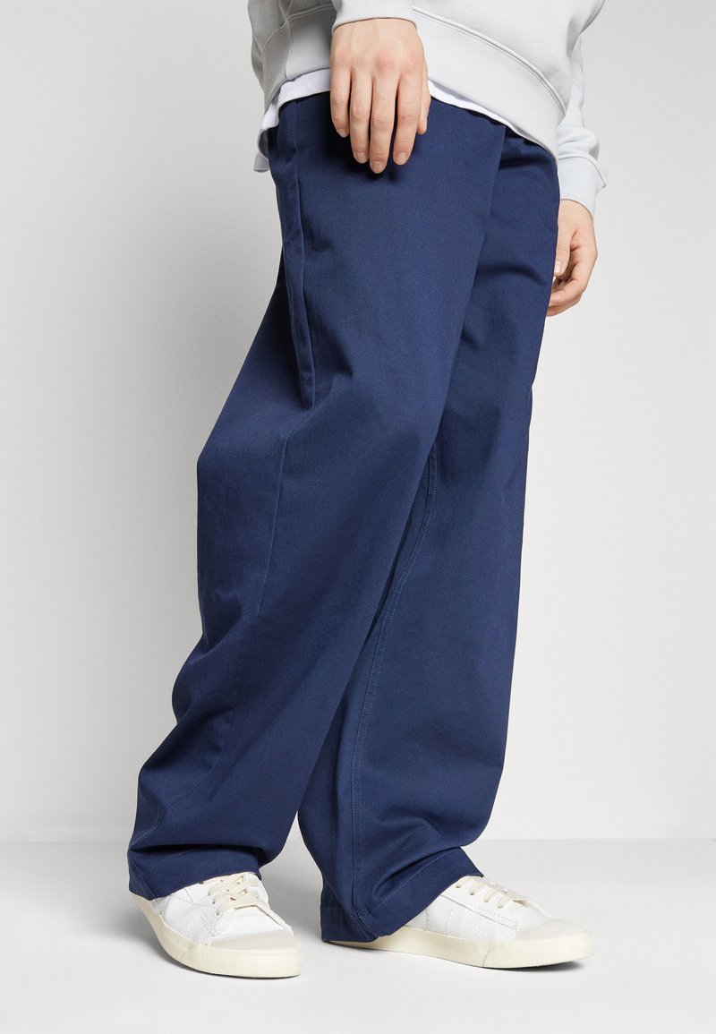 Pantaloni a gamba larga blu navy realizzati in tessuto di cotone, dotati di ampie tasche laterali e una vestibilità rilassata, abbinati a sneakers bianche.