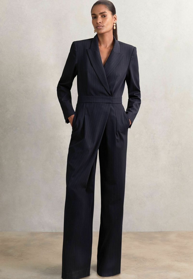 Donna che indossa una tuta blu navy a righe con pantaloni larghi, top in stile blazer e mani nei taschini, su uno sfondo neutro.