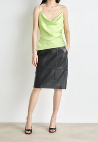 Grünes Satin-Camisole-Top mit dünnen Trägern, kombiniert mit einem schwarzen Bleistiftrock aus Leder mit Nahtdetails und Taschen. Schwarze Sandalen mit offenem Zehenbereich.