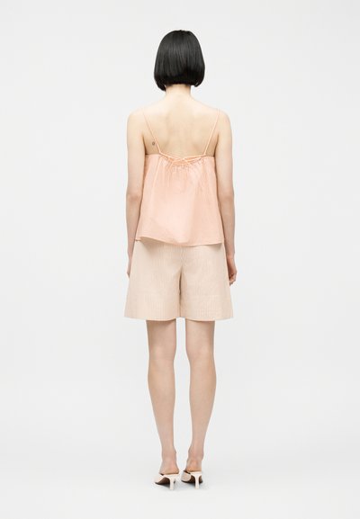 Γυναίκα με κοντά μαύρα μαλλιά στέκεται με την πλάτη, φοράει τοπ χωρίς μανίκια με ροδακινί και λευκές ρίγες και ταιριαστά σορτς μέχρι το γόνατο με ψηλοτάκουνα παπούτσια.