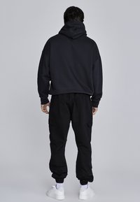 Sudadera negra con capucha de corte relajado, combinada con pantalones cargo negros con bolsillos laterales y zapatillas blancas sobre un fondo sencillo.