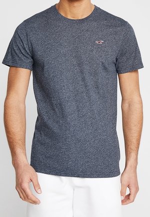 Mann trägt ein eng anliegendes, kurzärmeliges, dunkelgraues T-Shirt mit kleinem roten und weißen Logo auf der linken Brust, kombiniert mit weißen Shorts.