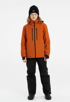 Orangewasserdichte Jacke mit Kapuze, Frontreißverschluss und zwei Brusttaschen. Kombiniert mit schwarzen Hosen und einer schwarzen gestrickten Mütze.