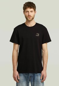 Camiseta de algodón negra con mangas cortas, cuello redondo y un pequeño parche de logo en el pecho izquierdo. Combinada con unos vaqueros de denim de color azul claro.