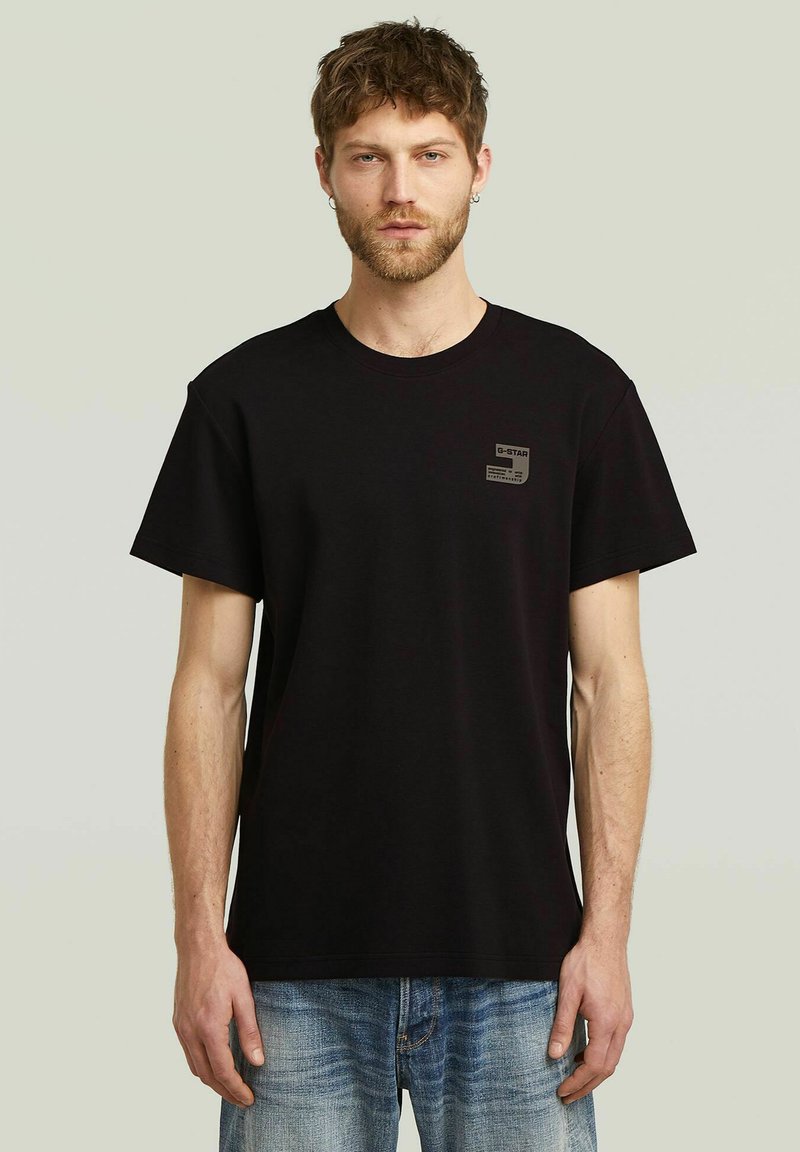 Camiseta de algodón negra con mangas cortas, cuello redondo y un pequeño parche de logo en el pecho izquierdo. Combinada con unos vaqueros de denim de color azul claro.