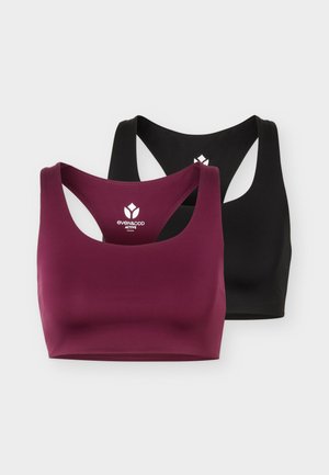 Slank persoon staand rechtop met een bordeauxrode sportbeha, zwarte cropped leggings, witte sokken en zwarte sportschoenen tegen een effen achtergrond.